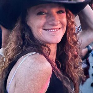 young woman with long curly brown hair and a friendly smile . She is wearing a cowboy hat and a tank top with one shoulder turned toward the camera.